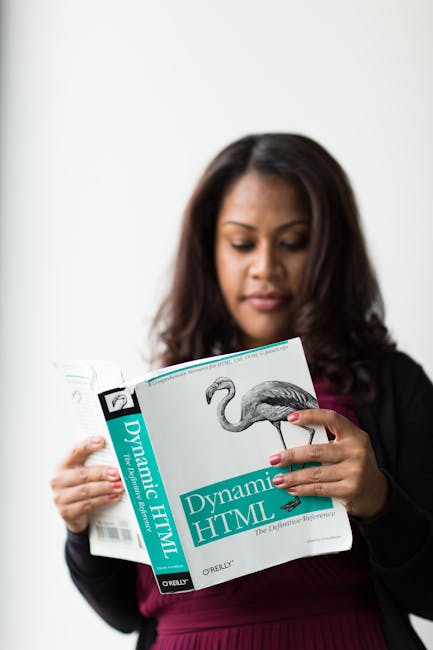 A woman with medium brown skin and long, dark hair is standing indoors against a plain white wall. She is wearing a black blazer over a dark red or burgundy top and is holding an open book titled 'Dyn