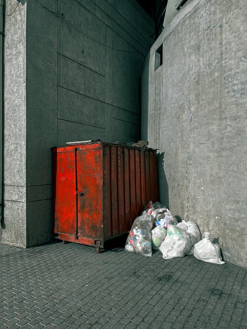 A row of dark grey wheelie bins is positioned along the sidewalk outside a red brick residential building, with each bin featuring a slightly open lid and white graffiti markings on their surfaces. A