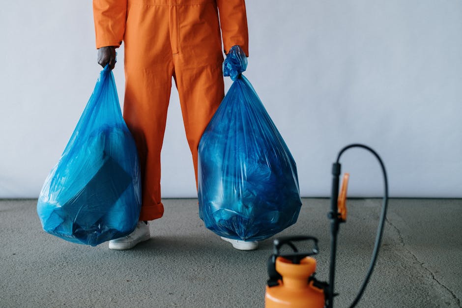 A close-up of multiple black plastic garbage bags filled with waste, tightly knotted at the top, positioned in a pile against a plain, light-colored wall. The bags have a shiny, crinkled texture, indi
